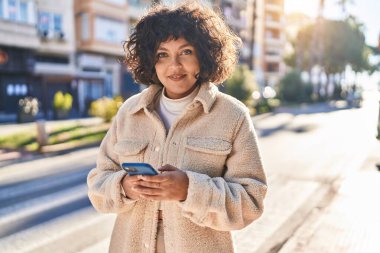 Genç, güzel, İspanyol bir kadın sokakta akıllı telefon kullanarak gülümsüyor.