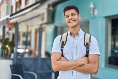 Genç İspanyol öğrenci gülümsüyor. Kollarını kavuşturup caddede duruyor.