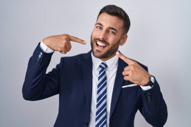 Yakışıklı İspanyol takım elbiseli ve kravatlı neşeli gülümseyen ve parmaklarıyla ve ağzıyla işaret eden adam. diş sağlığı kavramı. 