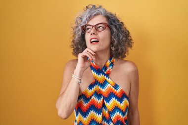 Middle age woman with grey hair standing over yellow background with hand on chin thinking about question, pensive expression. smiling with thoughtful face. doubt concept. 