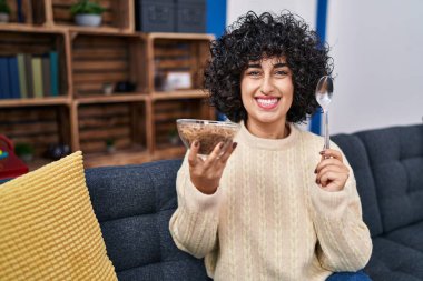 Kıvırcık saçlı genç esmer kadın sağlıklı tahıl gevrekleri yiyor ve yüzünde mutlu bir gülümsemeyle gülümsüyor. diş gösterme. 