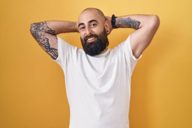 Sakallı, dövmeli, sarı arka planda duran, kolları ve elleri başının arkasında olan, mutlu gülümseyen genç İspanyol bir adam. 