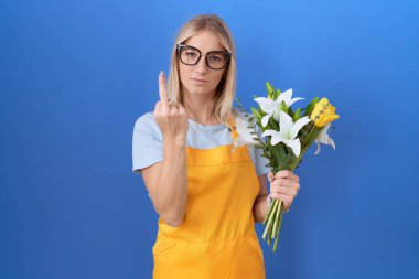 Young caucasian woman wearing florist apron holding flowers showing middle finger, impolite and rude fuck off expression 