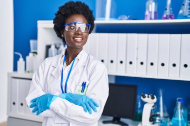 Genç Afrikalı Amerikalı bilim kadını laboratuvarda kollarını kavuşturup el kol hareketi yapıyor.