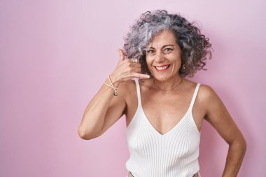 Middle age woman with grey hair standing over pink background smiling doing phone gesture with hand and fingers like talking on the telephone. communicating concepts. 
