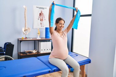 Genç Latin kadın hamile hasta klinikte lastik bant kullanarak fizyoterapi seansı yapıyor.