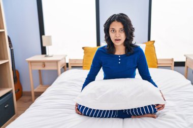 Evdeki yatakta oturan koyu renk saçlı, ağzı açık, korkmuş ve şaşırmış bir kadın. 