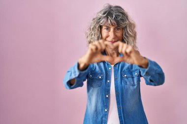 Pembe arka planda duran orta yaşlı bir kadın. Sevgiyle gülümseyen, elleriyle kalp sembolü yapan. romantik kavram. 