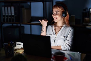 Young caucasian woman working at the office at night smiling with happy face looking and pointing to the side with thumb up. 