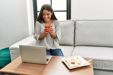 Genç Latin kadın kahvaltısını yapıyor. Evdeki kanepede oturuyor.