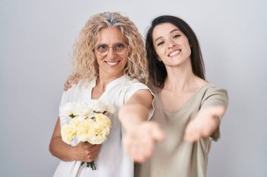 Anne ve kızı ellerinde bir buket beyaz çiçek tutarken mutlu bir gülümsemeyle avuç içi yardım ve kabullenme sunuyorlar.. 