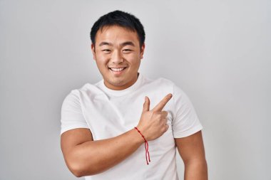 Genç Çinli bir adam beyaz arka planda neşeli bir şekilde ayakta duruyor. Yüzünde gülümseme var ve mutlu ve doğal bir ifadeyle elini ve parmağını kaldırıyor. 