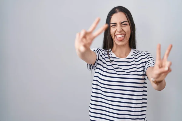 縞模様のTシャツを着た若いブルネットの女性が勝利サインをして両手の指を見せて舌で笑っている — ストック写真