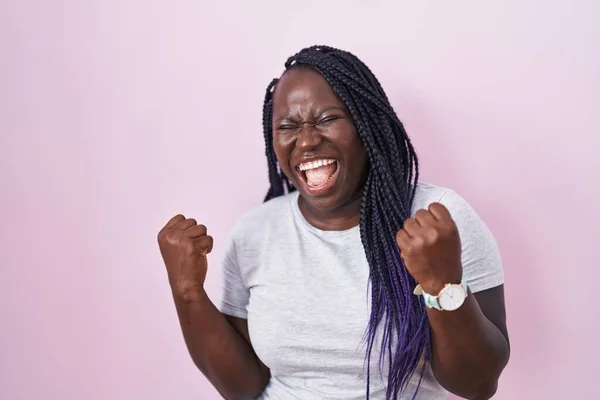 ピンクの背景に立っている若いアフリカの女性は非常に満足し 腕を上げて勝者のジェスチャーを行う興奮し 笑顔と成功のために叫んでいます お祝いのコンセプト — ストック写真