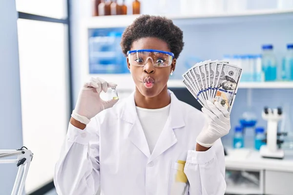 African american woman working at scientist laboratory holding dollars making fish face with mouth and squinting eyes, crazy and comical.