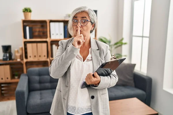 stock image Middle age woman with grey hair at consultation office asking to be quiet with finger on lips. silence and secret concept. 