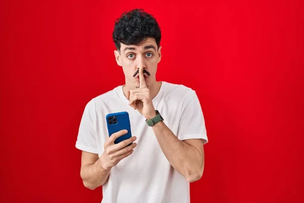 Hispanic Man Using Smartphone Red Background Asking Quiet Finger Lips — Stock Photo, Image
