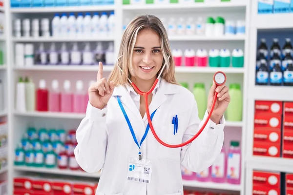 在药店工作的金发年轻女子拿着听诊器笑着 满脸喜形于色地指指点点 位居第一 — 图库照片
