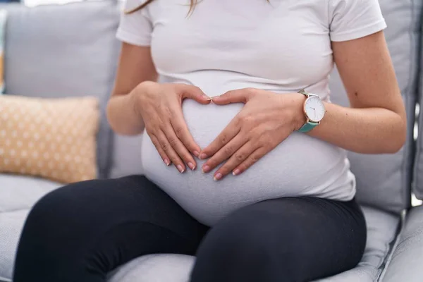 Young pregnant woman doing heart gesture on belly sitting on sofa at home