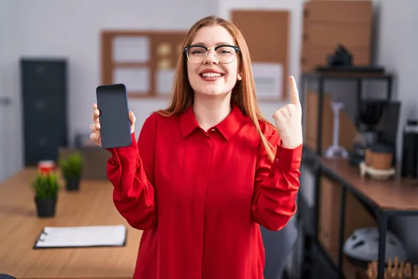 オフィスで働く赤毛の女性は 幸せな顔をしたアイデアや質問を指差す指で笑顔でスマートフォンの画面を示しています ナンバーワン — ストック写真