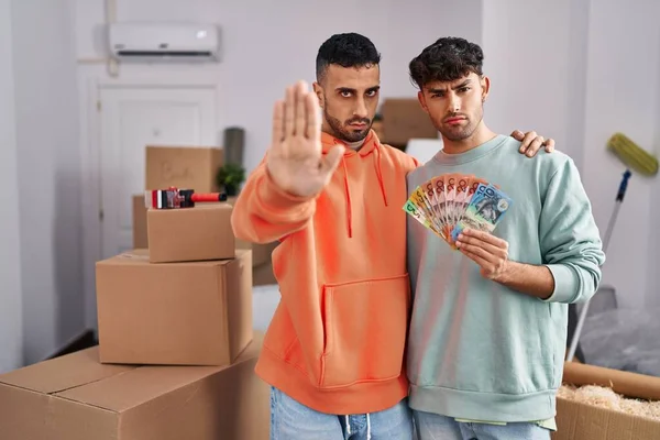 Young hispanic gay couple moving to a new home holding banknotes with open hand doing stop sign with serious and confident expression, defense gesture