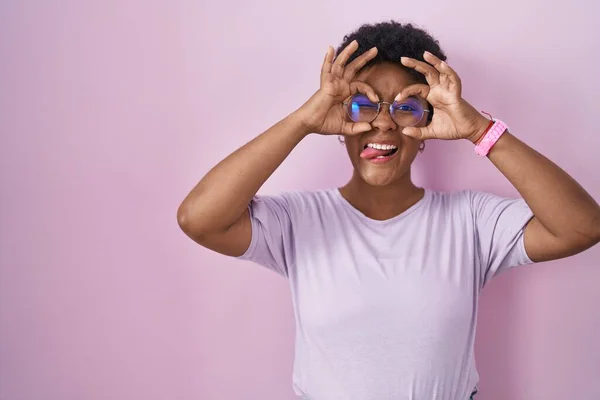 ピンクの背景の上に立つ若いアフリカ系アメリカ人の女性は 双眼鏡が舌を突き出しているようなOkジェスチャーをして 目は指を通して見る クレイジーな表現 — ストック写真