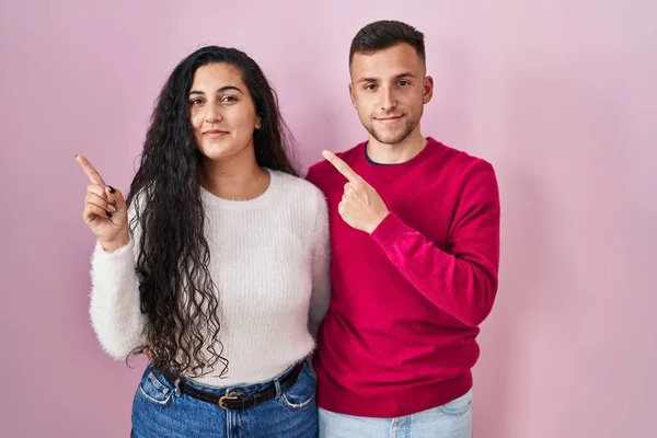 Unga Latinamerikanska Par Står Över Rosa Bakgrund Pekar Med Handen — Stockfoto