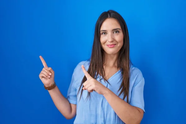 年轻的黑发女人笑着站在蓝色的背景上 看着摄像机 用双手和手指指向旁边 — 图库照片