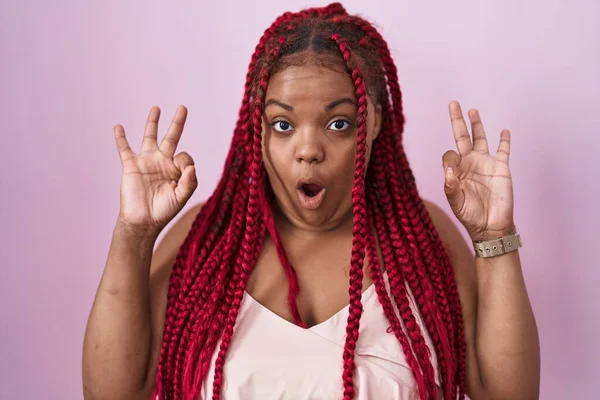 身穿辫子的非裔美国妇女站在粉色背景上 看上去惊讶而震惊 她用手指做了一个很好的认可标志 疯狂的表达 — 图库照片