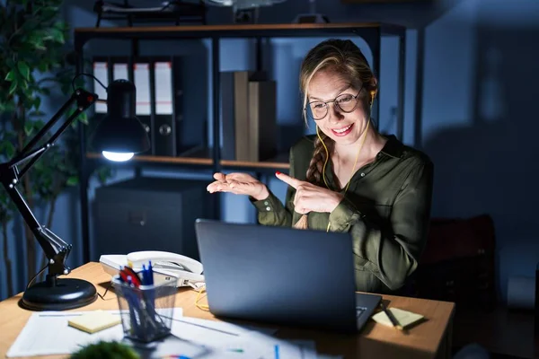 stock image Young blonde woman working at the office at night amazed and smiling to the camera while presenting with hand and pointing with finger. 