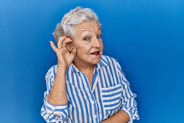 噂やゴシップに耳を傾ける耳の上に手で笑みを浮かべて青の背景に立って灰色の髪を持つシニア女性 聴覚障害の概念 — ストック写真