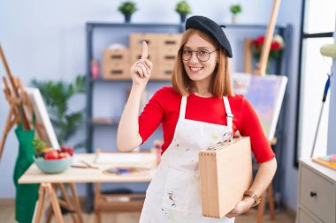 Sanat stüdyosundaki genç kızıl saçlı kadın elinde sanat çantası tutuyordu. Şaşırmış, şaşırmış ve parmaklarıyla işaret ediyor ve kollarını kaldırıyordu.. 