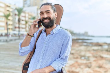Young hispanic man musician talking on smartphone holding guitar case at seaside