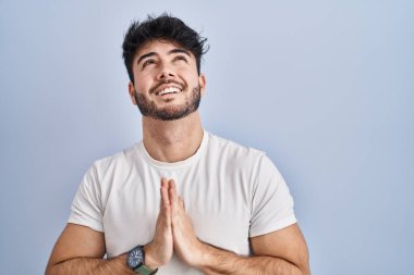 Hispanic man with beard standing over white background begging and praying with hands together with hope expression on face very emotional and worried. begging. 