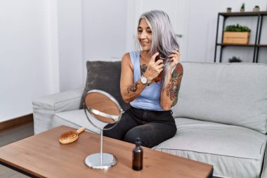 Orta yaşlı, kır saçlı bir kadın evdeki kanepede oturup saç bakımını yaptırıyor.