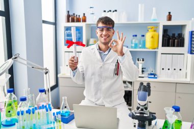 Laboratuvarda çalışan Latin asıllı genç bir adam diplomasını tutuyor. Parmaklarıyla imza atıyor, gülümsüyor ve mükemmel bir sembol. 