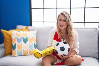 Young blonde woman supporting soccer team sitting on sofa at home