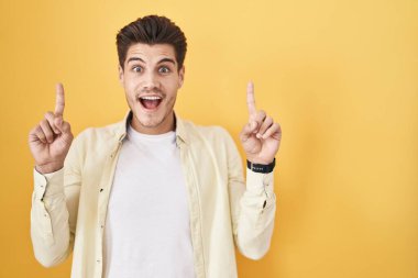 Young hispanic man standing over yellow background smiling amazed and surprised and pointing up with fingers and raised arms. 