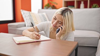 Not defterine yazı yazan genç, güzel İspanyol kadın evde akıllı telefondan konuşuyor.