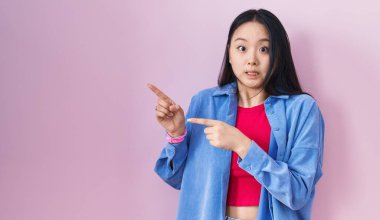 Young asian woman standing over pink background pointing aside worried and nervous with both hands, concerned and surprised expression 