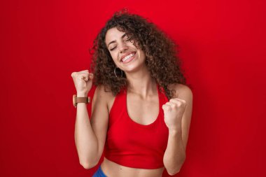 Kırmızı arka planda duran kıvırcık saçlı İspanyol kadın kolları havada ve gözleri kapalı bir şekilde başarıyı kutluyor ve şaşırıyor. Kazanan konsept. 