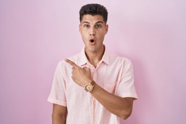 Young hispanic man standing over pink background surprised pointing with finger to the side, open mouth amazed expression. 