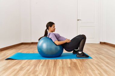 Spor merkezinde formda top kullanan genç Latin kadın karın kası egzersizi yapıyor.
