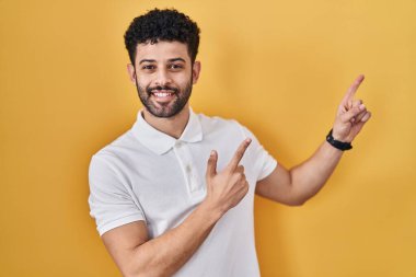 Arap adam sarı arka planda duruyor gülümsüyor ve kameraya bakıyor iki eliyle ve parmaklarıyla kenarı işaret ediyor.. 