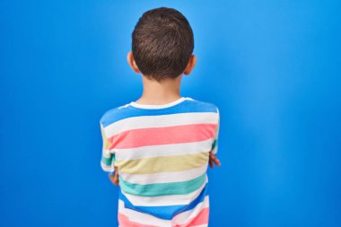 Genç beyaz çocuk mavi arka planda dikiliyor arkasını dönüp kollarını kavuşturmuş bakıyor. 