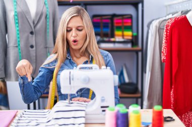Sarışın kadın terzi, dikiş makinesi kullanıyor. Parmaklarıyla işaret ediyor. Reklam veriyor, şaşırıyor ve ağzını açıyor. 