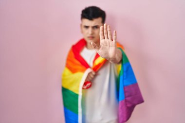 İkili olmayan kişi elinde gökkuşağı lgbtq bayrağı, elinde açık elle dur işareti ve ciddi ve kendinden emin bir ifadeyle savunma hareketi yapıyor. 