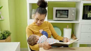 Anne ve oğul yemek odasında bebek emzirirken kitap okuyorlar.