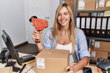 Young blonde woman ecommerce business worker holding packing tape machine at office
