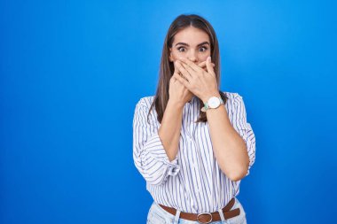 Hispanic young woman standing over blue background shocked covering mouth with hands for mistake. secret concept. 